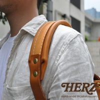 職人手作皮革復古風旅行後背包，展示其棕褐色皮革肩帶，帶有縫線和兩顆黃銅鉚釘，搭配淺色亞麻襯衫。