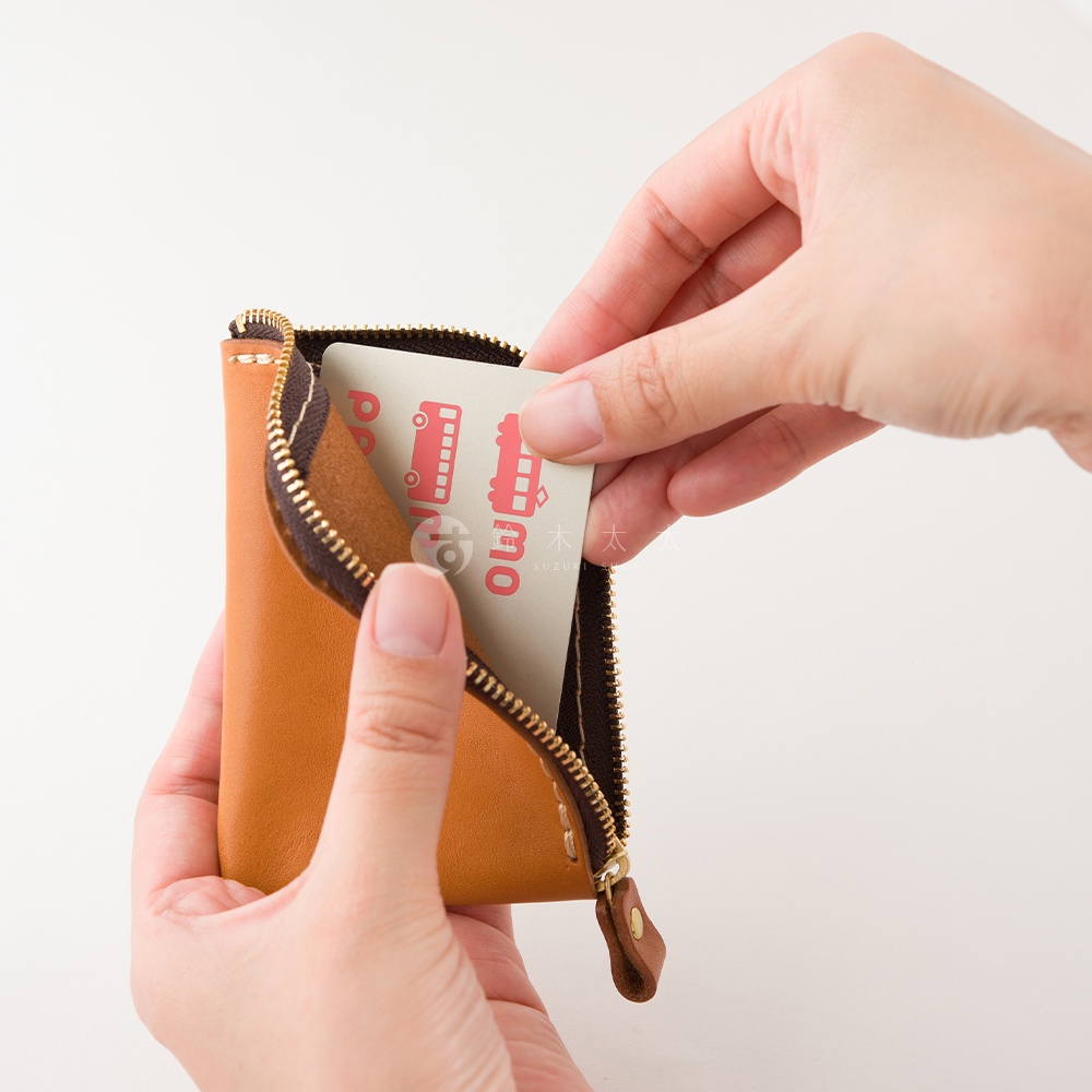 雙手持拿一個日本HERZ職人手作的棕色皮革迷你零錢包，拉鍊呈現L型開口，一隻手正將一張印有粉紅色巴士圖案的卡片放入錢包夾層中，展示其小巧精緻的收納設計。