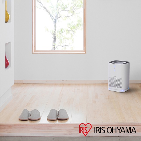 白色簡約風格情境式空氣清淨機 IAP-A25，方形機身，頂部有控制面板，前側有網狀進氣孔，底部有藍色指示燈，放置於木質地板上，背景有窗戶及綠色植物。