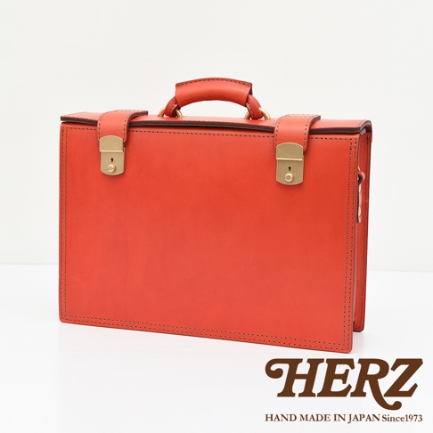 橘紅色皮革製成的手提箱，配有金色鎖扣、提把和側邊金屬細節。