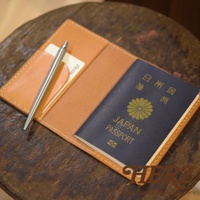 職人手作皮革經典護照套，淺棕色皮革，內含一本深藍色日本護照和一支銀色金屬筆，置於木質桌面。