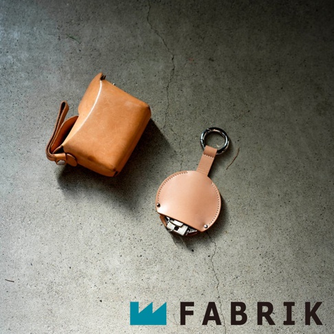 FABRIK 皮革圓形收納鑰匙圈和方形皮夾，兩種皮革製品一同置於水泥地表面，品牌 LOGO 位於圖片右下角。