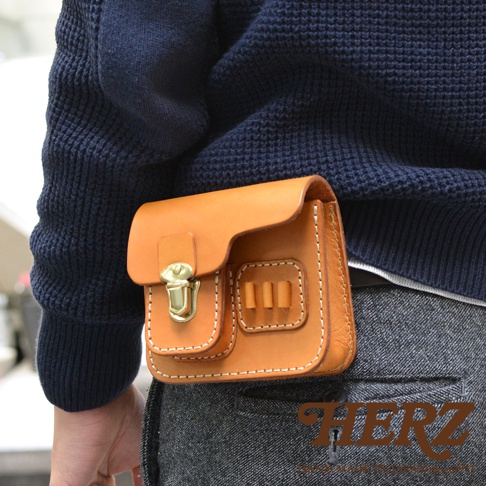 職人手作咖啡色皮革高爾夫腰包，款式簡約，設有金屬鎖扣及可插置物品的隔層，搭配深藍色針織衫及灰色褲子。