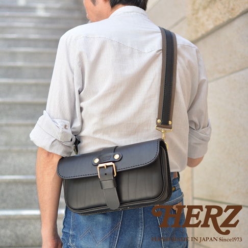 黑色職人手作皮革單扣經典斜背包，配有織帶和黃銅色金屬扣環，由皮革製成，斜挎於身穿條紋襯衫的男士肩上。