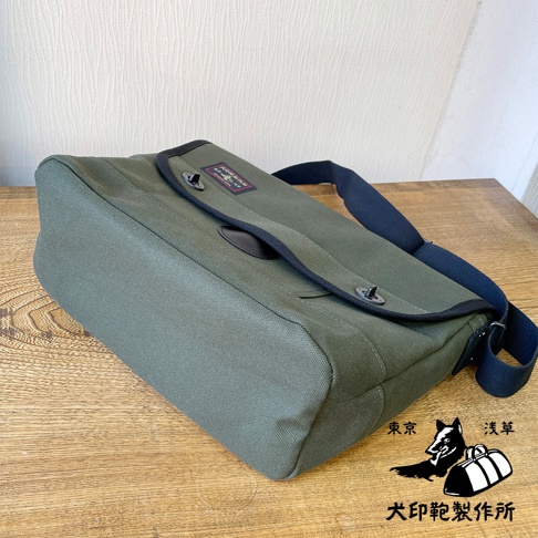 軍綠色純棉帆布雙扣郵差包，配有黑色織帶，包身方形，側面有明顯的布料紋理。