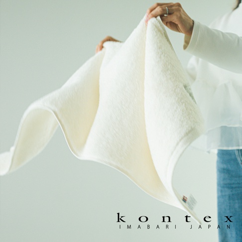 一位女性雙手舉起一條柔軟的米白色 KONTEX 品牌大毛巾，展示其細緻的毛圈質感與輕盈垂墜感，背景為簡約的室內空間。