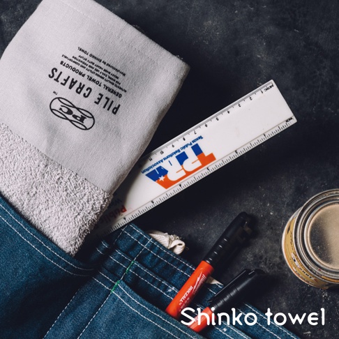 素面極簡風格的淺灰色毛巾，帶有黑色文字標誌 PILE CRAFTS 及 PC 圖案，毛巾下半部為白色毛圈，旁邊置有一支帶有 Texas Public Relations Association 標誌的白色尺、一支紅色簽字筆及一個金屬罐。