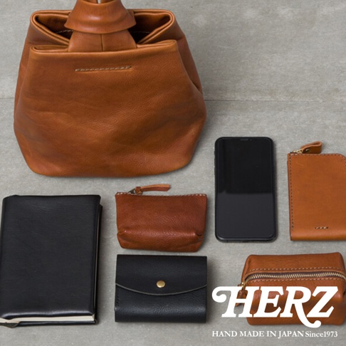 職人手作皮革Organ單柄手提包，呈現棕色。圖片中另有黑色筆記本、棕色零錢包、黑色短夾、棕色拉鍊小包和黑色手機。