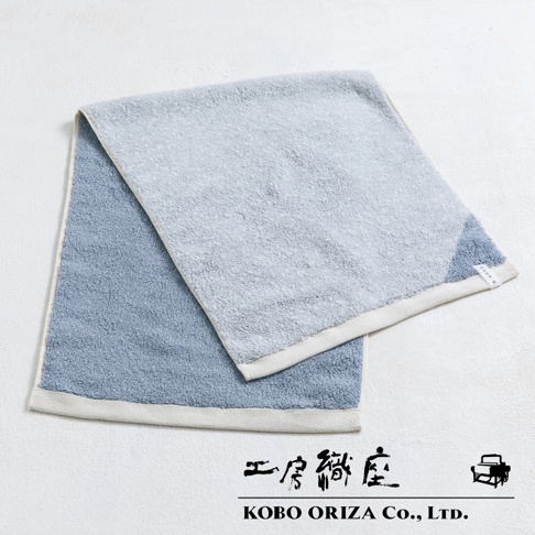 一條棉麻混紡雙色快乾毛巾，一邊為淺藍色，另一邊為淺灰色，兩色交界處有藍色拼接，毛巾邊緣為米白色滾邊。
