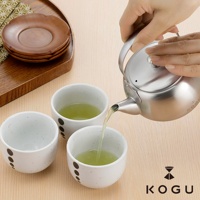 日製18-8不鏽鋼急須茶壺，壺身為銀色，附有濾網，正由手持注入綠茶，旁邊擺放著三個裝有綠茶的白色陶瓷茶杯，茶杯上有黑色圓點裝飾。