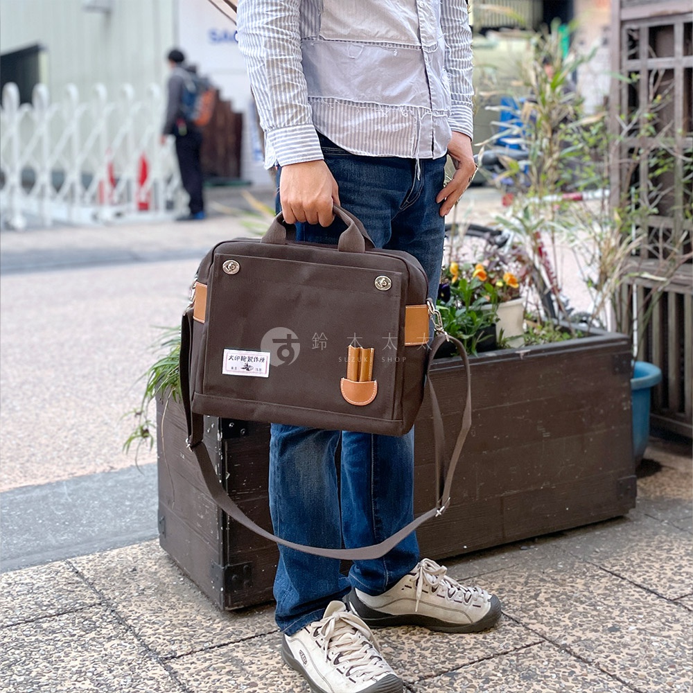 咖啡色純棉帆布製成的橫式三用側背包，配有棕色皮革裝飾與提把，側面有掛繩，適合日常攜帶。