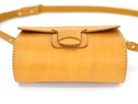 職人手作皮革寬扣帶馬鞍包，呈現溫潤的淺棕色調。包身為圓筒狀，採對比色縫線。包蓋處設有皮革製的寬版扣帶，搭配金屬釦環，簡潔而實用。