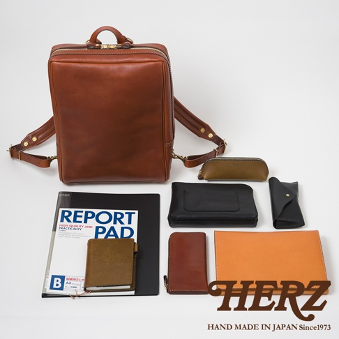 職人手作皮革軟皮商務後背包，搭配同款棕色皮革筆袋、黑色皮夾、黑色皮質平板電腦包、棕色皮革筆記本、橘色皮革文件夾，以及一份標示「REPORT PAD」的報告。