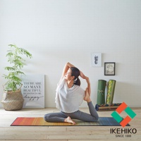 一位女性在色彩漸層的藺草榻榻米瑜珈墊上做著瑜珈動作。墊子為大地色系，搭配綠色及紅色交織的logo。背景有植物、裝飾畫及捲起的瑜珈墊。