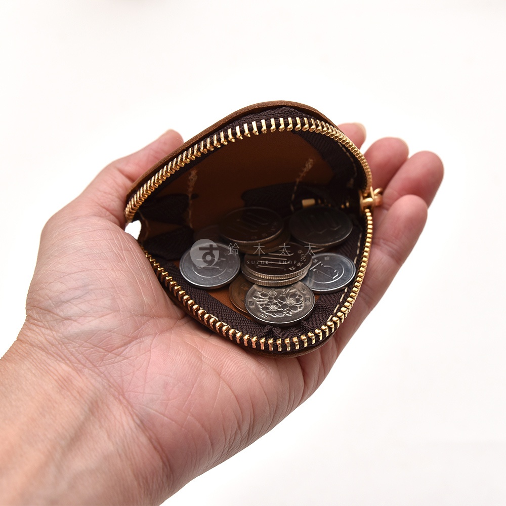 一隻手拿著日本HERZ職人手作皮革三角飯糰造型零錢包，皮質呈現溫潤的棕色，拉鍊開口處露出內部裝載的多枚硬幣，展現出精緻且實用的生活感。