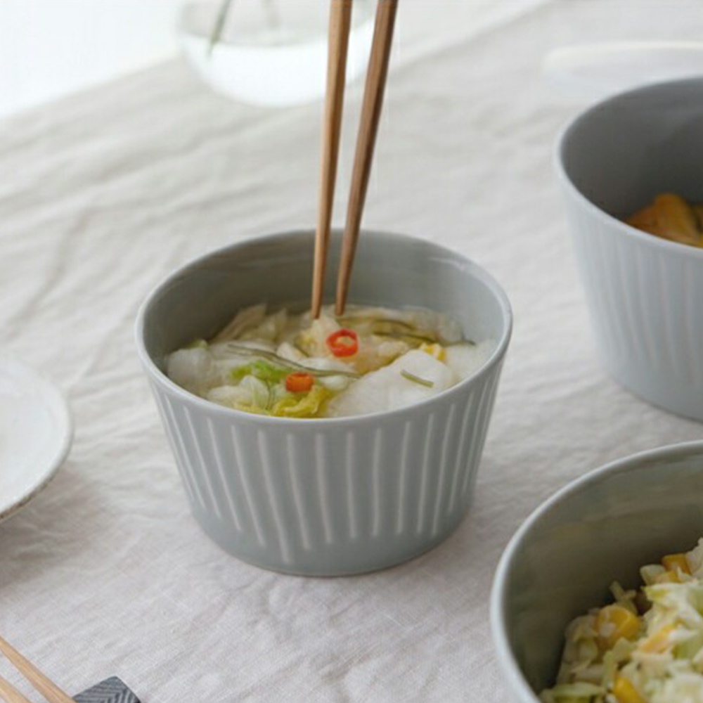 淺灰色陶瓷微波小碗，碗身帶有垂直細紋，內盛醃漬白菜，有紅辣椒點綴，一雙木筷夾取碗中食物。