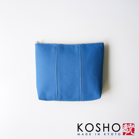 一個京都KOSHO品牌的藍色帆布化妝收納包，外觀呈現長方形，表面有三條垂直的車縫線裝飾，右下角印有KOSHO MADE IN KYOTO的黑色文字與紅色方形印章圖案。