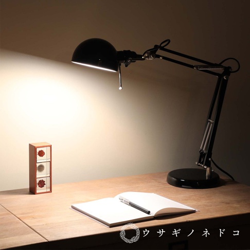 木質三層小收納櫃，木紋淺棕色，每層櫃門有紅色花朵圖案，置於木桌上，旁為黑色可調式檯燈，檯燈下有打開的筆記本和筆。