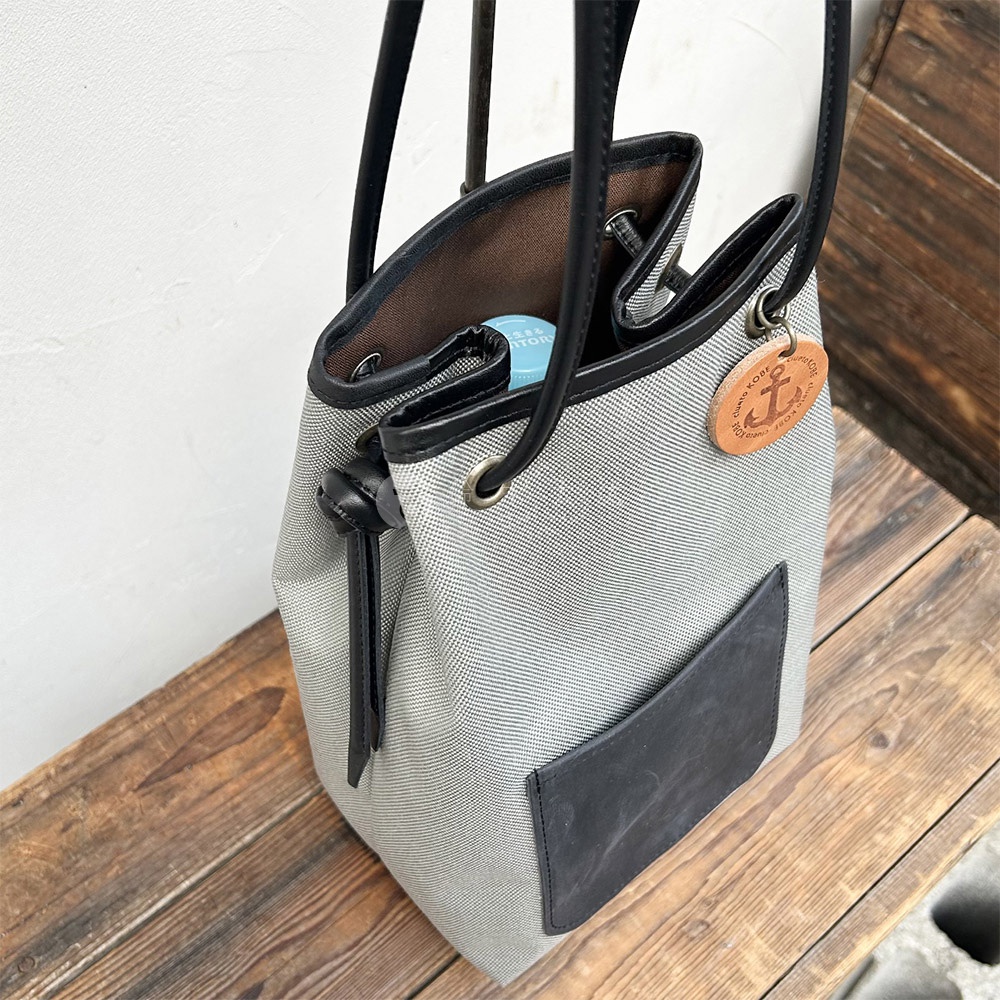 左側斜視圖，展示一個灰色防潑水尼龍束口袋肩背包，配有義大利皮革口袋和一個船錨圖案的吊牌。