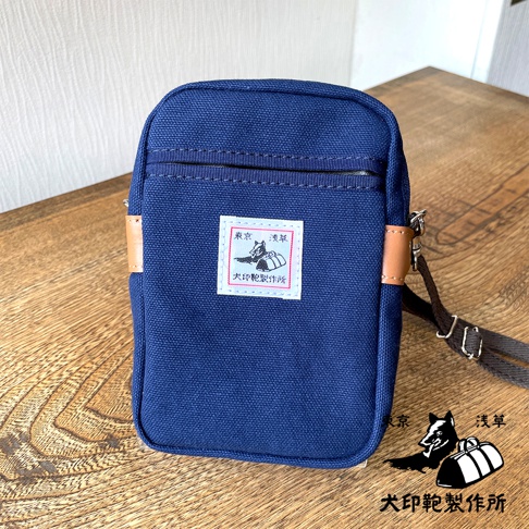石洗帆布斜背手機包，藍色，配有皮革飾邊和可調節斜背帶，包身中央有方形布貼，印有日文圖案和文字。