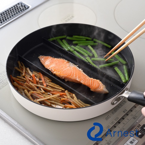 一個白色的 Arnes 三合一分隔不沾平底鍋放置在電磁爐上，鍋內由隔板分為三個區域，分別正在烹煮煎鮭魚、炒牛蒡胡蘿蔔絲以及綠色蔬菜，一雙木筷正夾取鍋內的綠色蔬菜，鍋身呈現現代簡約風格。