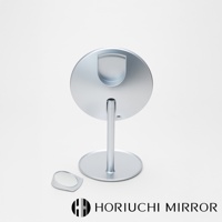 銀色金屬材質的桌上型化妝鏡，圓形鏡面，帶有支架和底座，鏡背有半圓形置物槽，旁邊放著一個小型三倍放大鏡。