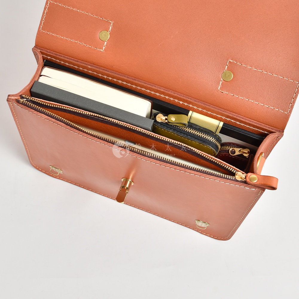 開放式棕色皮革公事包，內部有筆記本、手機、綠色與紅棕色零錢包，並以拉鍊與銅扣固定。