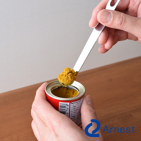 一隻手拿著不鏽鋼萬用醬料小勺，勺頭盛著黃色粉末，準備舀入紅色罐中。