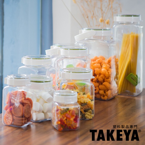 多個透明塑膠密封收納罐，其中裝有義大利麵、小糖果、棉花糖、餅乾、通心粉等，罐子皆有白色蓋子和綠色密封圈，擺放在木質桌面。