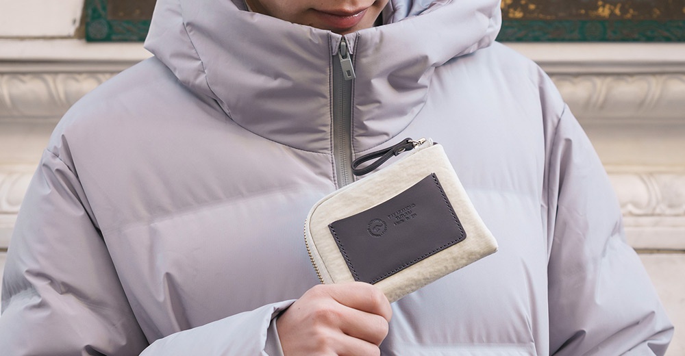 一個人在粉紫色的羽絨外套內，手持一個淺色、帶有深棕色皮革標籤的拉鍊零錢包。