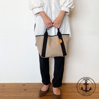 米色帆布托特包，配有黑色織帶裝飾和黑色皮革提把，包上掛有圓形皮革吊牌，風格簡約。