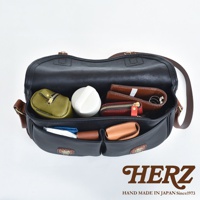 黑色皮革傳統釣魚包，內部裝有綠色零錢包、白色圓形物品、紅色拉鍊零錢包、兩件棕色皮夾及其他小物。包款有黑色主體和棕色皮革提把及肩帶，前方有兩個翻蓋式口袋。