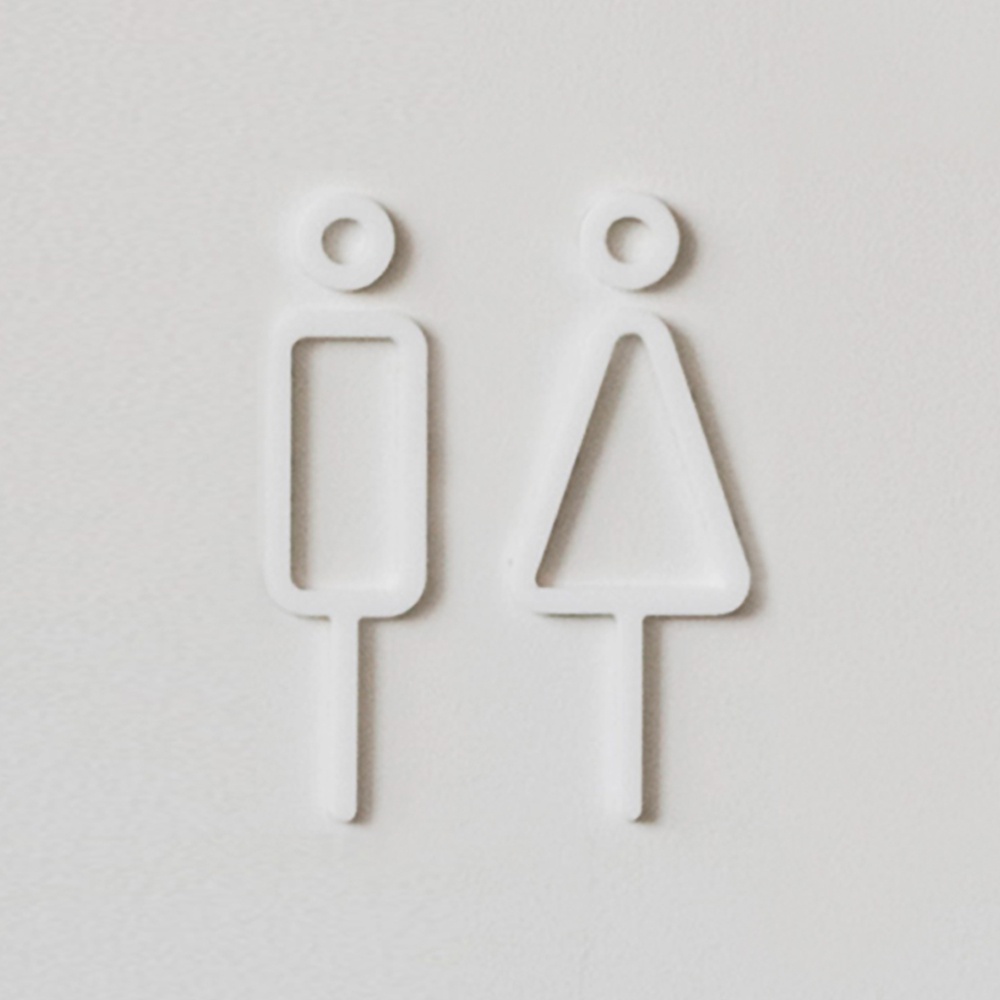 白色簡約風格的男女廁所標示牌，左邊為男性圖示（長方形身體），右邊為女性圖示（三角形身體），皆配有圓形頭部及下方指示棒，材質疑似為壓克力或金屬。