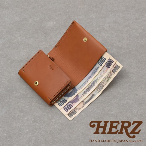 一款日本HERZ手工製作的棕色皮革迷你短夾，以俯視視角呈現，錢包為雙折式設計並印有Organ字樣，內部夾層展示三張不同面額的日圓紙鈔，整體風格簡約俐落，展現出皮革細膩的質感與職人手作工藝。