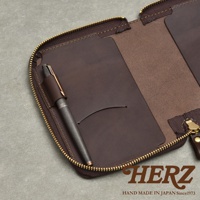 一個深棕色皮革記事本收納包，內有筆套插著一支金屬筆，角落有HERZ品牌標誌和HAND MADE IN JAPAN Since1973字樣。