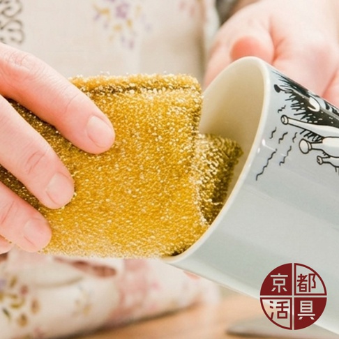 一雙手正拿著一塊金色顆粒質感的京都活具菜瓜布，正在清洗一個繪有卡通圖案的馬克杯，畫面下方帶有京都活具的圓形品牌標誌。
