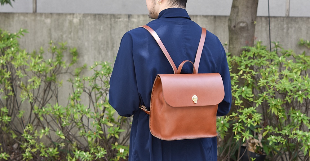 一款棕色皮革翻蓋迷你後背包，配有同色系皮革肩帶和金色鎖扣，由一位穿著海軍藍上衣的人背負。