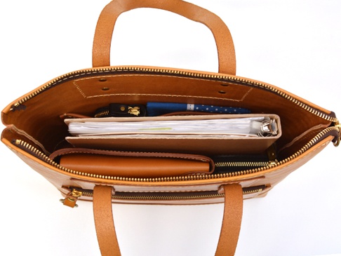 職人手作皮革威靈頓直式拉鍊托特包，呈現溫潤的棕褐色，內部可見文件夾、筆記本、筆和零錢包。包體採用天然皮革，質感厚實，並配有金屬拉鍊及提把，設計簡約實用。