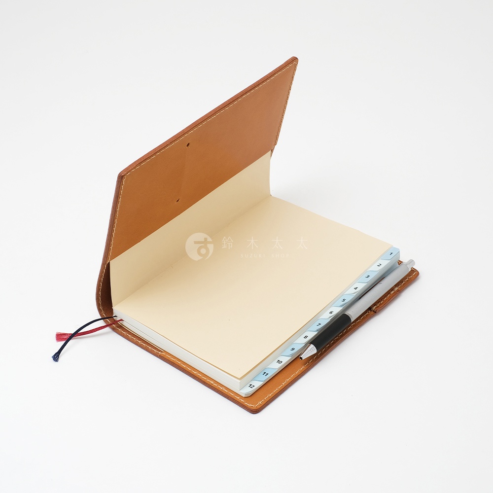 職人手作皮革B6記事本書套，展示為淺棕色皮革，內頁為米白色，附有標示數字1至12的索引分隔頁，書套夾有黑色筆及紅色與藍色書籤繩。