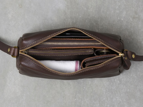 職人手作皮革Organ輕巧拉鍊單肩包 (G-80) M，呈現深棕色粒面皮革，包身拉鍊敞開，內部收納著手機、皮夾、零錢包和一條白色毛巾，搭配金色拉鍊與黃銅色金屬扣環，散發質感。