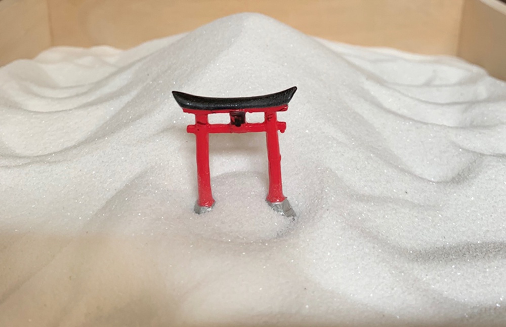 一個小型的紅色鳥居，頂部為黑色，立於白色細沙堆砌的枯山水庭園模型中。
