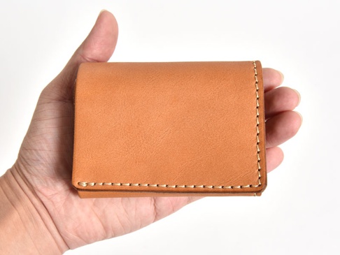 手持一個棕褐色皮革三折式迷你短夾，帶有明顯的縫線細節，簡潔的設計展現皮革的自然質感。