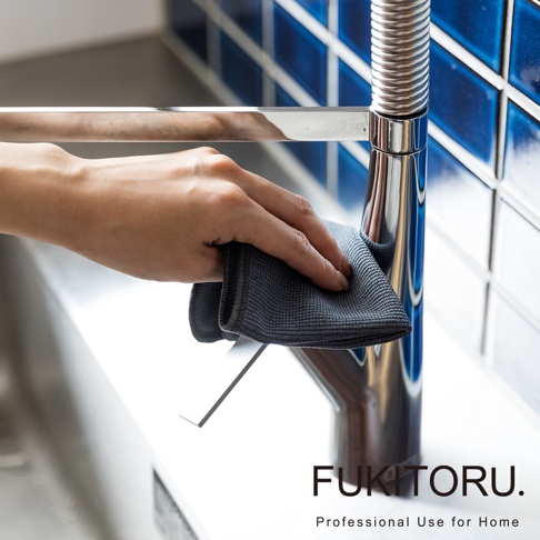 一隻手正拿著一塊深灰色超細纖維擦拭布，擦拭著閃亮的金屬水龍頭。背景是藍色的磁磚牆面。
