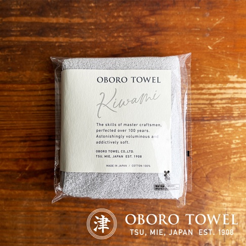 包裝內的灰色細纖維毛巾，表面有細緻的織紋，標籤上印有「OBORO TOWEL Kiwami」、「The skills of master craftsmen, perfected over 100 years. Astonishingly voluminous and addictively soft.」、「OBORO TOWEL CO.,LTD. TSU, MIE, JAPAN EST. 190