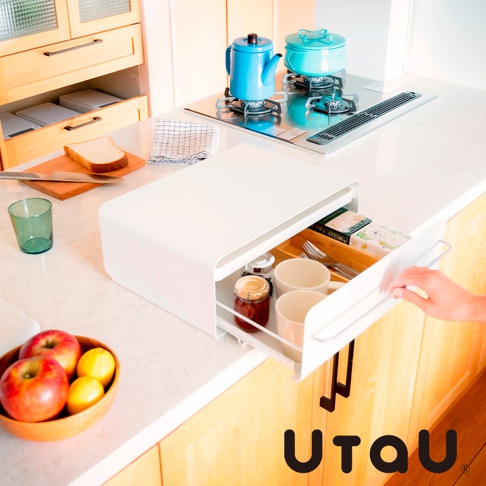 白色抽屜式收納櫃，櫃內有兩個馬克杯、一罐果醬和餐具。櫃子下方是木質櫥櫃，上面擺放著一個木碗裝著蘋果和檸檬，以及一個綠色玻璃杯。