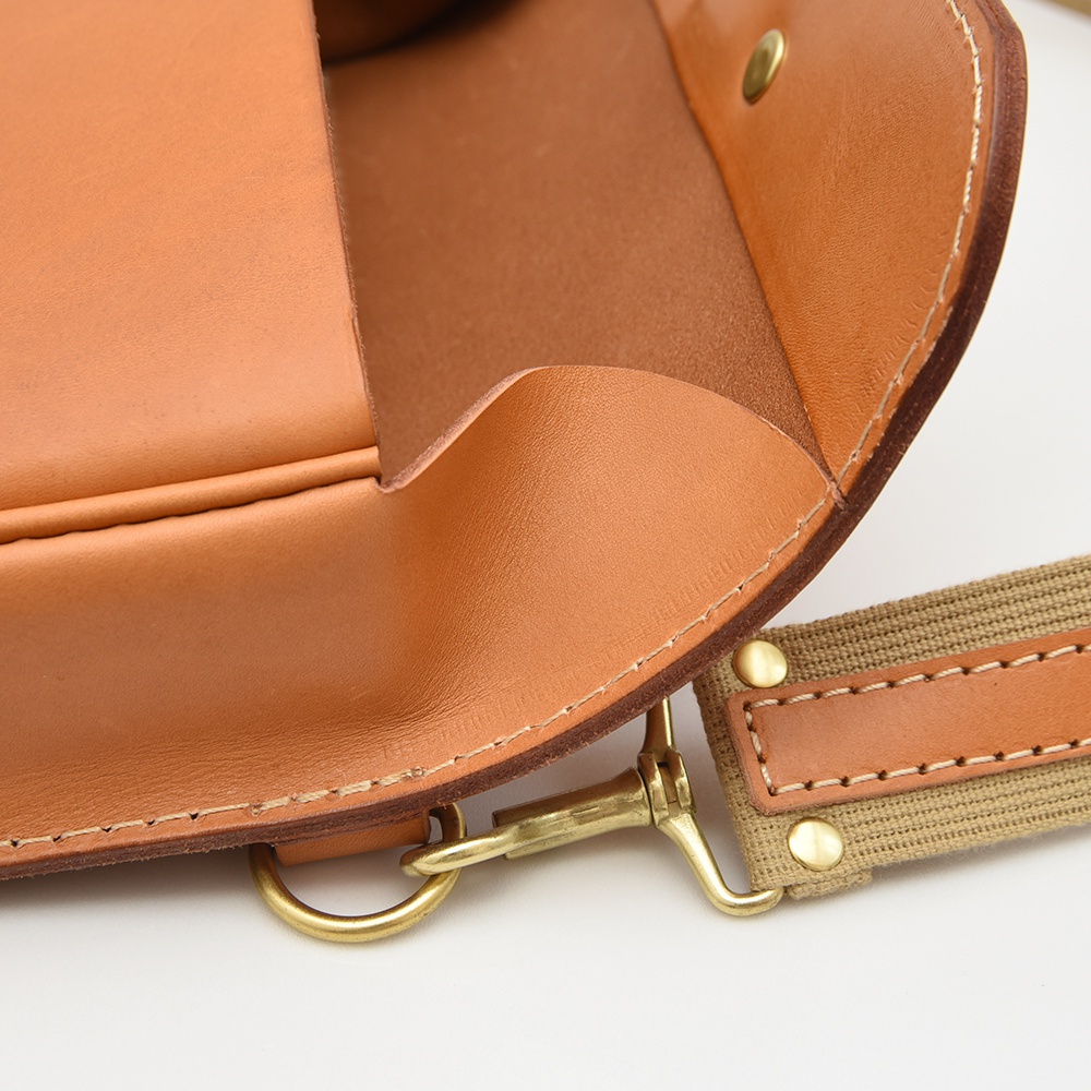 特寫：職人手作皮革單扣經典斜背包，主要為棕褐色光滑皮革材質，搭配帆布織帶和黃銅色金屬釦環。