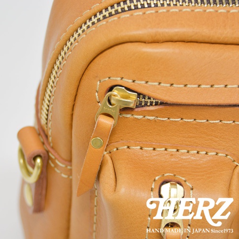 職人手作皮革單肩包，使用淺棕色皮革，搭配金色拉鍊和金屬配件，縫線明顯，質感良好，風格簡約。
