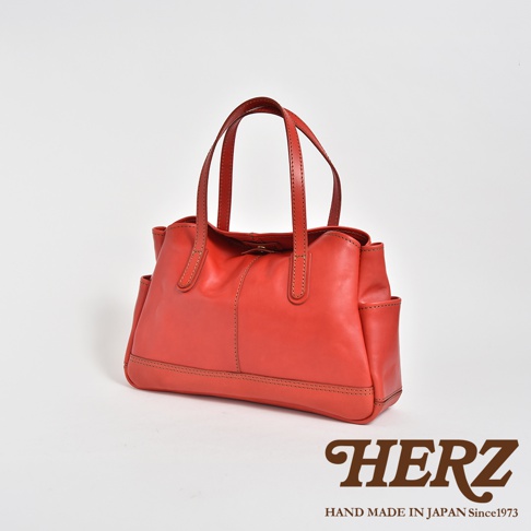 一款由日本HERZ品牌職人手工製作的紅色皮革托特包，包身設計簡約，採用高品質皮革製成，側面設有實用口袋，底部縫線細膩整齊，搭配兩條堅固的皮革提把，整體呈現出經典且耐用的手作質感。