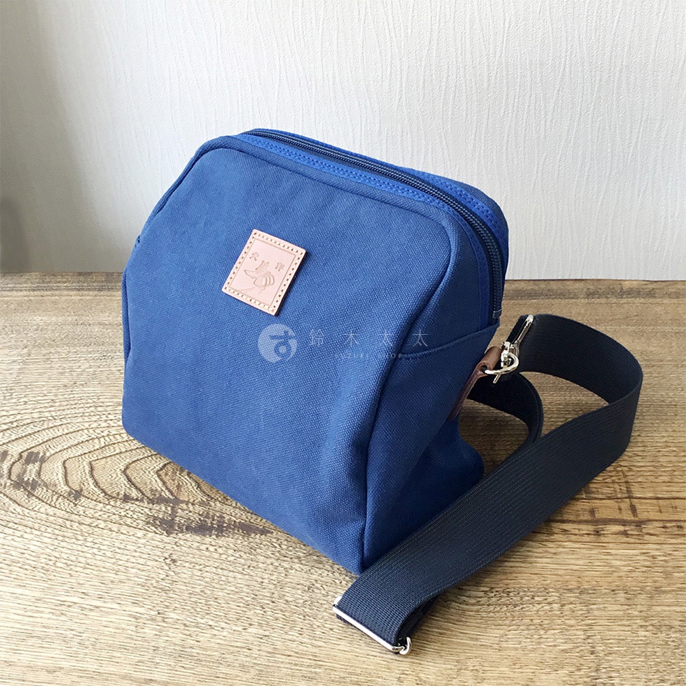 藍色石洗帆布材質散步包，正面有方形皮革標籤，印有小蜜蜂圖案及品牌標誌，附有黑色織帶斜背包帶。