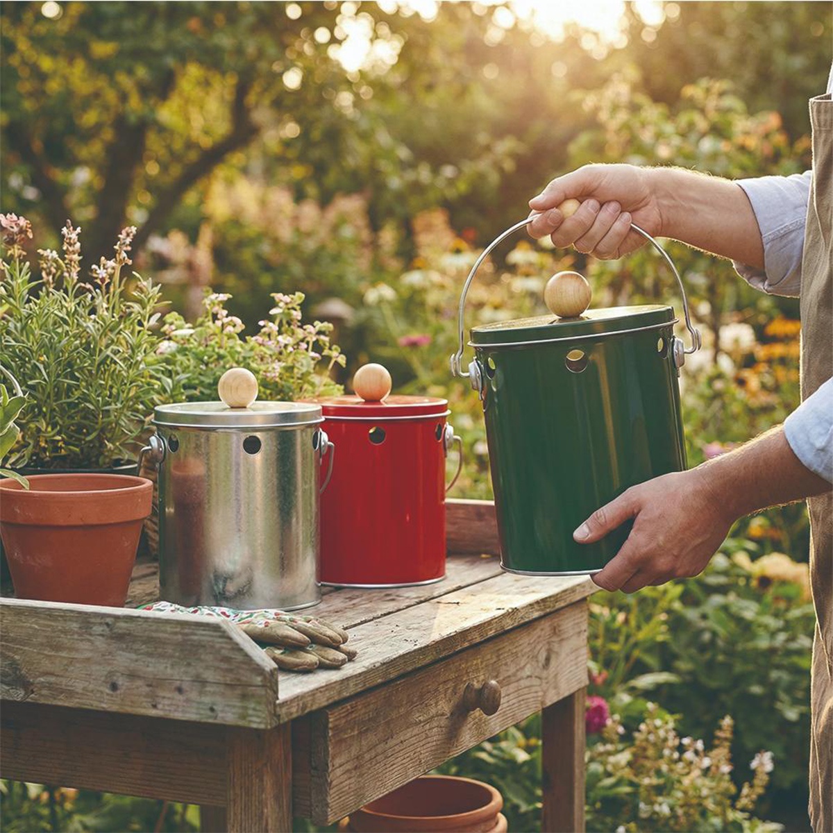 男人在花園的木桌旁，拿著綠色的 OBAKETSU 蚊香桶，旁邊還有一個銀色和一個紅色的蚊香桶，以及陶盆和園藝手套。
